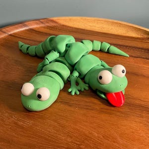 May include: A green articulated lizard toy with large white eyes and a red tongue. The toy is made of plastic and is lying on a wooden surface. The lizard's body is segmented, allowing for flexible movement.