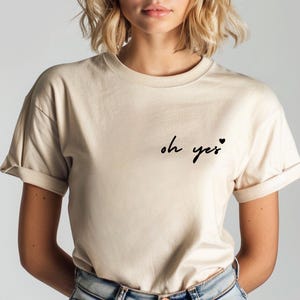 Puede incluir: Una camiseta beige con el texto "oh yes" y un peque&ntilde;o coraz&oacute;n impreso en la parte delantera.