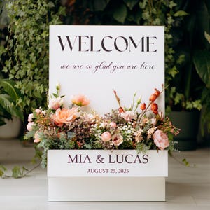 May include: A white welcome sign with the text "WELCOME" and "we are so glad you are here" in a script font. The sign is decorated with a floral arrangement of peach and pink flowers. The names "MIA & LUCAS" and the date "AUGUST 25, 2025" are also displayed.