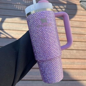 May include: A purple, bedazzled Stanley tumbler with a matching handle. The tumbler has a white lid with a pink top and a silver band. The Stanley logo is visible on the front of the tumbler. The tumbler is held against a wooden background.