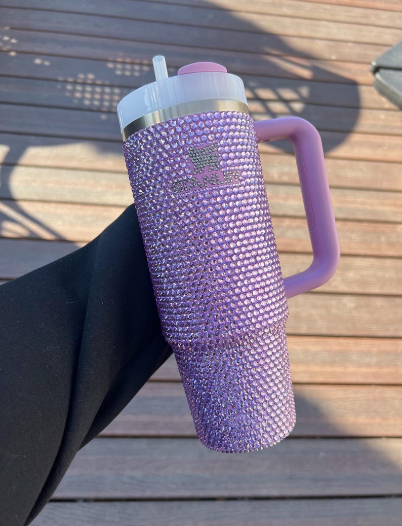 May include: A purple, bedazzled Stanley tumbler with a matching handle. The tumbler has a white lid with a pink top and a silver band. The Stanley logo is visible on the front of the tumbler. The tumbler is held against a wooden background.
