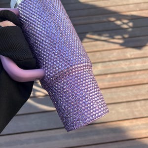 May include: A lavender tumbler covered in small, sparkling rhinestones. The tumbler has a white rim and a clear straw. The handle is a light purple color. The tumbler is held against a wooden background.