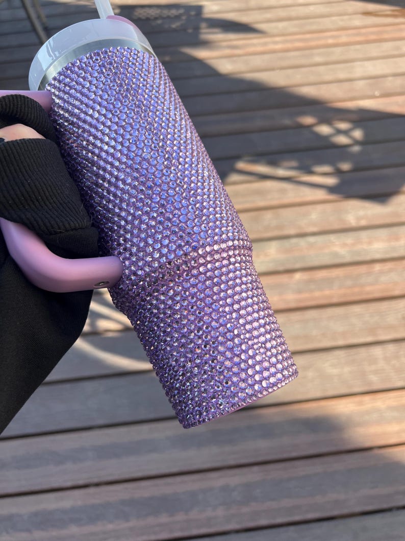 May include: A lavender tumbler covered in small, sparkling rhinestones. The tumbler has a white rim and a clear straw. The handle is a light purple color. The tumbler is held against a wooden background.