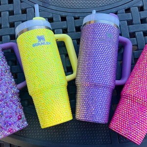 May include: Four Stanley tumblers in vibrant colors: purple, yellow, and pink. The purple and pink tumblers are covered in sparkling rhinestones. The yellow tumbler features the Stanley logo. Each tumbler has a handle and a straw.