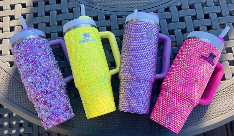 May include: Four Stanley tumblers in vibrant colors: purple, yellow, and pink. The purple and pink tumblers are covered in sparkling rhinestones. The yellow tumbler features the Stanley logo. Each tumbler has a handle and a straw.