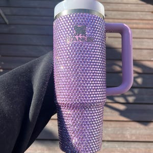 May include: A purple, bedazzled Stanley mug with a handle. The mug is covered in small, sparkling rhinestones. It has a white rim and a light purple handle and lid. The Stanley logo is visible on the front of the mug.