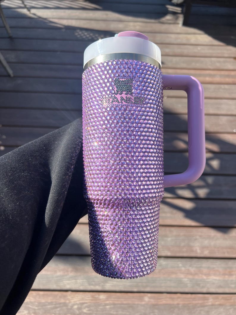 May include: A purple, bedazzled Stanley mug with a handle. The mug is covered in small, sparkling rhinestones. It has a white rim and a light purple handle and lid. The Stanley logo is visible on the front of the mug.