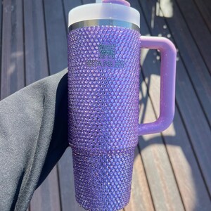 May include: A purple tumbler covered in sparkling rhinestones. The tumbler has a handle and a lid with a straw. The Stanley logo is visible on the side. The handle and lid are a lighter shade of purple.
