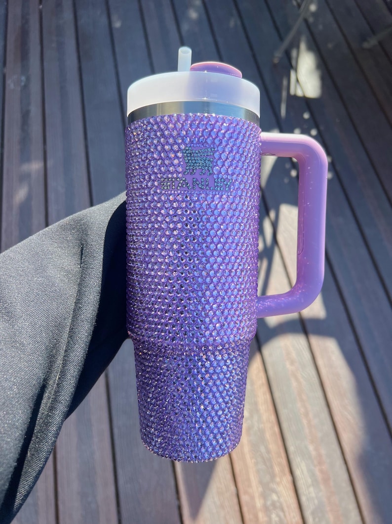 May include: A purple tumbler covered in sparkling rhinestones. The tumbler has a handle and a lid with a straw. The Stanley logo is visible on the side. The handle and lid are a lighter shade of purple.