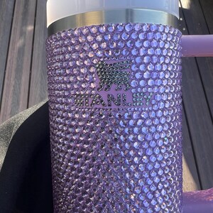 May include: A purple Stanley tumbler covered in sparkling rhinestones. The tumbler has a white lid, a silver band, and a purple handle. The Stanley logo is visible on the front of the tumbler.