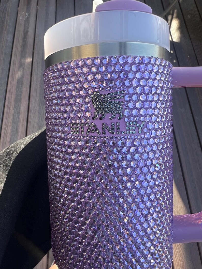 May include: A purple Stanley tumbler covered in sparkling rhinestones. The tumbler has a white lid, a silver band, and a purple handle. The Stanley logo is visible on the front of the tumbler.