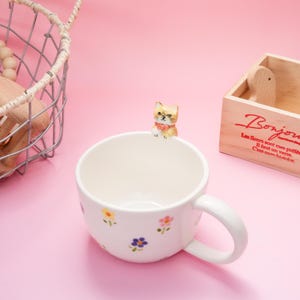 May include: A white ceramic mug with a handle, adorned with colorful floral patterns. A small, decorative cat figurine sits on the rim. The mug is on a pink surface, with a wooden box and wire basket in the background.