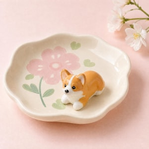 May include: A cream-colored ceramic dish shaped like a flower petal, featuring a painted pink flower and green leaves. A small, three-dimensional ceramic figurine of a brown and white Corgi dog is positioned in the center of the dish.