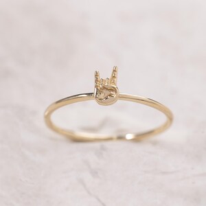 May include: A thin gold ring featuring a small, detailed charm in the shape of a hand making the rock and roll sign. The charm is centered on the top of the smooth, circular band, which is crafted from a polished, reflective gold-toned metal material.