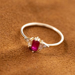 May include: A gold ring featuring a central rectangular red gemstone flanked by two smaller rectangular clear stones and two tiny round clear stones on each side, set in a thin polished gold band with a prong setting for each stone.
