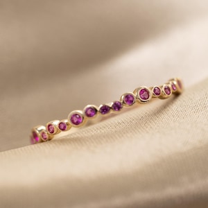 May include: A gold band ring featuring a series of small, round, bezel-set pink gemstones arranged in a row. The stones vary slightly in size and are set within a polished gold metal frame that follows the circular shape of the ring band.