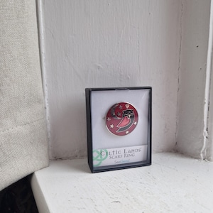 May include: A red and black enamel scarf ring featuring an owl sleeping on a crescent moon. The ring is presented in a small box with the words "Celtic Lands Scarf Ring" printed on it.