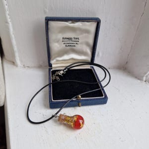 May include: A red glass vial filled with gold flakes and red flowers hangs from a black cord. The vial is attached to a silver chain and is in a blue velvet box with the text "Alexander Scott Jewellers & Silversmiths 102 Buchanan St. GLASGOW."