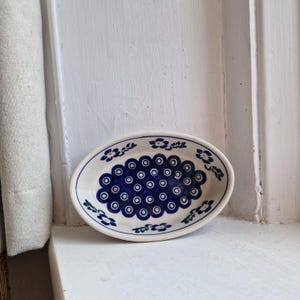 May include: Oval ceramic dish with a white base and a blue floral pattern. The dish features a central design of blue circles with white centres, surrounded by a border of smaller floral motifs. The dish is placed on a white surface.
