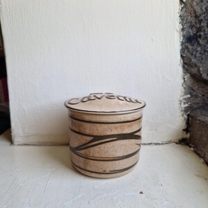 May include: A cylindrical ceramic jar with a matching lid featuring the word "caveau" in dark script. The jar has a speckled beige finish with horizontal and diagonal dark brown lines wrapping around the body, creating a textured appearance.