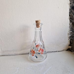 May include: A clear glass bottle with a narrow neck and a cork stopper. The body of the bottle features a hand-painted design of orange flowers with green leaves. The glass has a smooth, transparent finish and a rounded base shape.