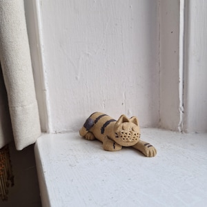 May include: A tan ceramic cat figurine with black stripes and a purple patch on its back. The cat is lying down with its paws extended. The figurine has a carved face and is resting on a white surface.