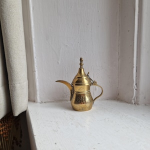 May include: A small, decorative brass teapot with a curved spout, handle, and hinged lid. The teapot has engraved floral patterns and a decorative finial. It is a vintage piece, likely used for serving coffee or tea.