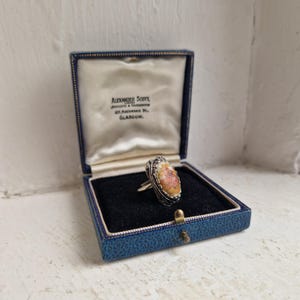 May include: An oval ring with a silver band and a central stone in pink, orange, and cream hues. The stone is set in a decorative silver filigree. The ring is presented in a vintage blue jewellery box with the text "ALEXANDER SCOTT" and "GLASGOW".