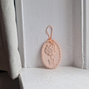 May include: A peach-coloured, oval-shaped ornament featuring a tree design. The tree is in a pot, and the ornament has a decorative border. A peach-coloured ribbon is attached for hanging.