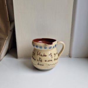 May include: A small cream-coloured ceramic pitcher with a brown interior and a single handle. The exterior features a hand-painted blue dotted rim and the handwritten text: Every blade of grass has its own drop of dew, with a wavy line underneath.