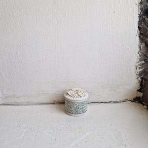May include: A small, cylindrical ceramic trinket box with a lid. The base features a dark green, abstract, linear pattern on a white background. The lid is adorned with a cluster of sculpted, three-dimensional white and pale yellow flowers.