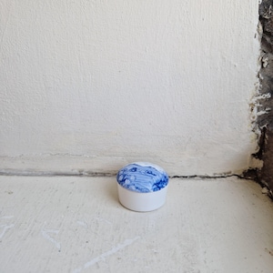 May include: A small, round, white ceramic container featuring a lid with a swirling blue and white marbled pattern. The base of the container is solid white, and the lid has a smooth, glossy finish with intricate, organic blue colour variations.