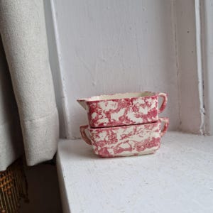 May include: Two stacked rectangular ceramic dishes with handles and a spout. The dishes have a cream base with a red marbled pattern. They sit on a white windowsill, with a curtain in the background. Each dish is approximately 15 cm long.
