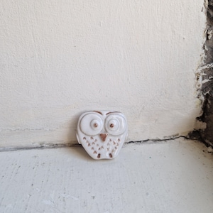 May include: A small, white ceramic owl figurine with a glossy finish. The owl features large, round eyes with brown pupils, a triangular brown beak, and a series of small, brown, V-shaped markings across its chest, representing feathers.