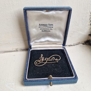 May include: A gold-toned metal brooch featuring the name Joan in cursive script, resting inside a blue hinged jewellery box. The interior lid is lined with white satin and printed with the text Alexander Scott, Limited, Jewellers & Silversmiths.