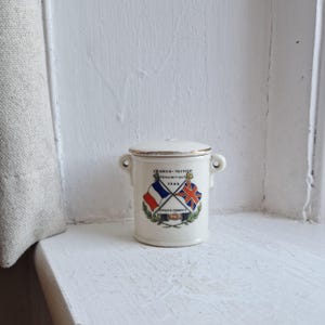 May include: A small, cream-coloured ceramic jar with a lid and two small handles. The jar features a design with the flags of France and the United Kingdom, crossed, with text that reads "FRANCO-BRITISH EXHIBITION 1908". The rim is trimmed with gold.