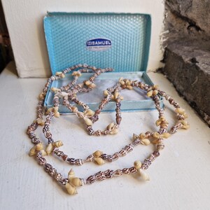 May include: A long necklace composed of small, spiraled brown and cream-coloured seashells strung together. The necklace is displayed in front of an open, light blue rectangular jewellery box with the text H. SAMUEL FOR FINE JEWELLERY inside.