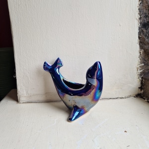 May include: A ceramic whale-shaped vase with a glossy, iridescent finish. The vase is mainly blue with hints of purple, green, and silver. It has a small opening for holding flowers or other decorative items. The whale's tail and head are distinct features.