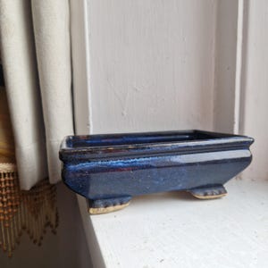 May include: A dark blue ceramic bonsai pot with a rectangular shape and a glossy finish. The pot has a flared design and rests on four small, rounded feet with a tan hue. The interior is a darker shade of blue.