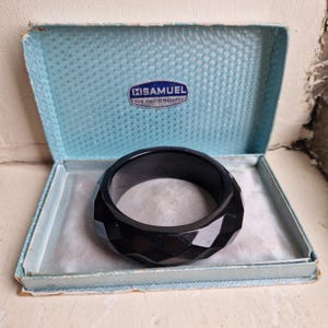 May include: A black faceted bangle bracelet resting inside a light blue hinged jewellery box. The interior lid of the box features a white label with the text H.SAMUEL FOR FINE JEWELLERY. The bracelet has a geometric, multi-sided surface.