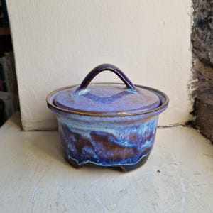 May include: A round ceramic lidded jar featuring a glossy glaze with blended shades of deep blue, purple, and light lavender. The lid has a curved handle, and the base of the jar is supported by small feet, showcasing a smooth, hand-crafted finish.