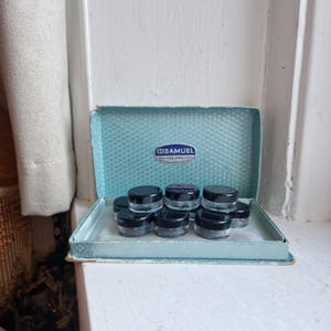 May include: A vintage jewellery box in light blue, containing several small, clear jars with black lids. The box has the text "SAMUEL FOR FINE JEWELLERS" on the inside. The box is open, revealing the jars inside.