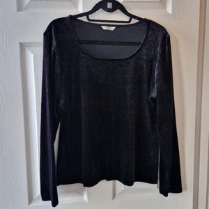 May include: A black velvet long-sleeved top with a scoop neckline hangs on a black hanger. The top has a soft, textured appearance and a simple, elegant design. The garment is displayed against a white door.