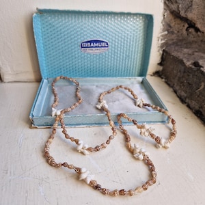 May include: A long necklace made of small brown spiral seashells interspersed with clusters of white shells. The necklace is displayed inside a light blue rectangular jewellery box with a textured lid featuring the text H.SAMUEL FOR FINE JEWELLERS.