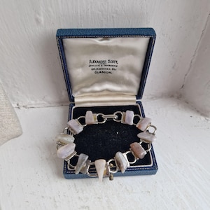 May include: A silver bracelet with a chain link design and a clasp. The bracelet is adorned with a series of light grey, brown, and white stones. The bracelet is in a blue velvet box with the inscription "Alexander Scott, Jewellers & Silversmiths, 100, Buchanan St., Glasgow."