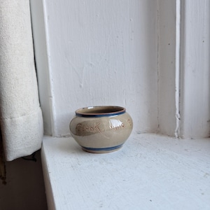 May include: A small, round, beige ceramic pot with a blue rim and a blue stripe near the base. The words "Speak Time" are etched into the side of the pot. The pot is sitting on a white surface.