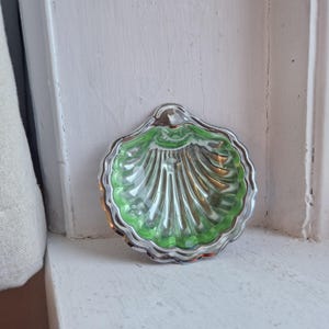 May include: A shell-shaped dish with a silver rim and a bright green interior. The dish features a detailed, ridged design. It is set against a white background. The dish is likely used for serving small portions or as a decorative item.