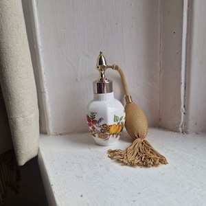 May include: A vintage white perfume bottle with a floral design in red, yellow, and green. It has a gold-coloured atomizer and a matching tassel. The bottle is likely made of porcelain.