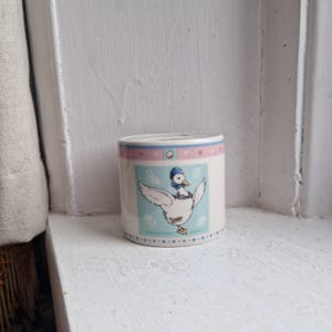 May include: A cylindrical ceramic container with a duck illustration. The container has a white base with a light blue panel featuring a cartoon duck wearing a blue hat and scarf. Pink and blue bands with polka dots and an egg design adorn the top.
