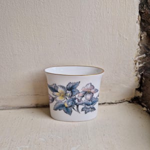May include: A white porcelain vessel with a flared rim and a gold-toned edge. The exterior features a botanical illustration of flowers with blue, yellow, and soft pink petals surrounded by dark blue-grey leaves arranged in a horizontal band.